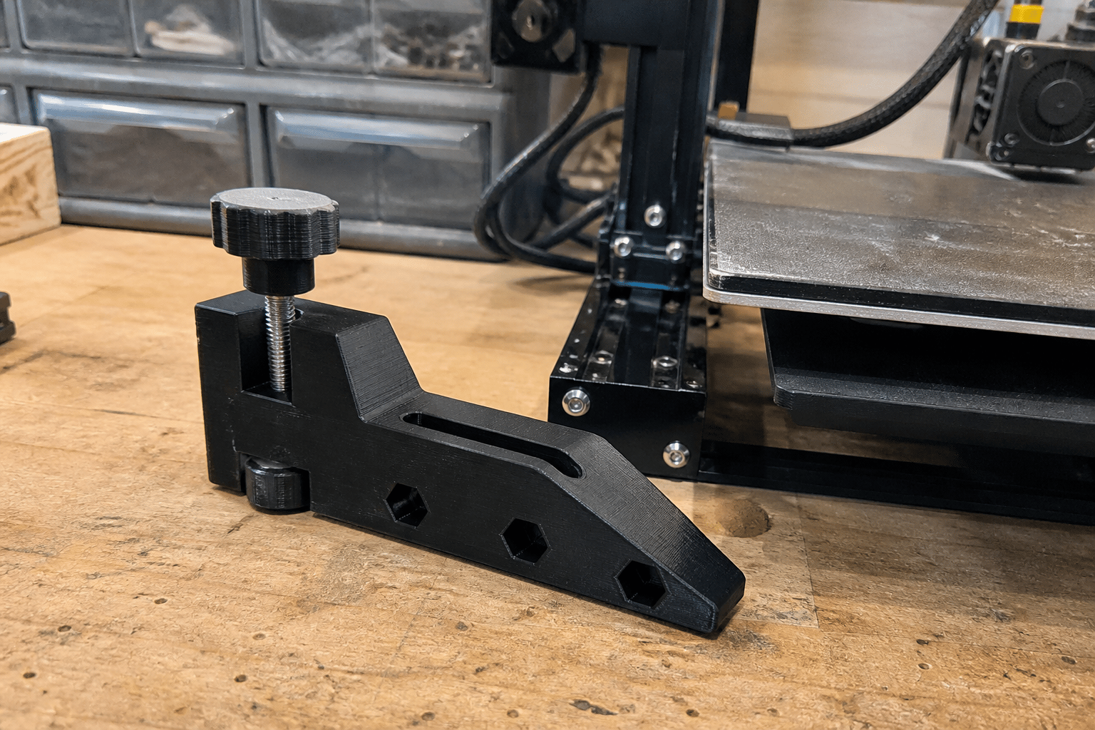 Black 3D-printed woodworking hold-down jig beside a printer
